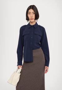 Navy blue shirt with a button-up front, two chest pockets, and shoulder epaulets, paired with a textured brown knit skirt and a white handbag.