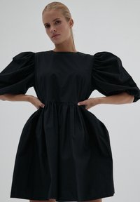 Blondynka z włosami związanymi w kok, ubrana w czarną sukienkę z bufiastymi, średniej długości rękawami, pozująca z dłońmi na biodrach na jednolitym tle.