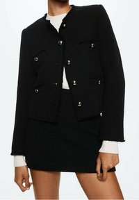 Veste noire texturée avec quincaillerie dorée, présentant un design court, quatre poches avant et un ourlet effiloché. Associée à une jupe noire.