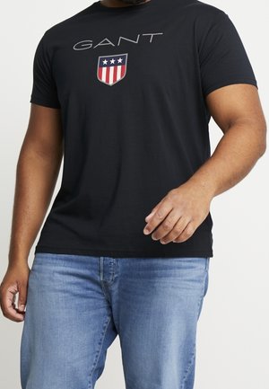 Hombre con camiseta negra de GANT con logo y vaqueros azules, de pie con una mano ligeramente levantada.