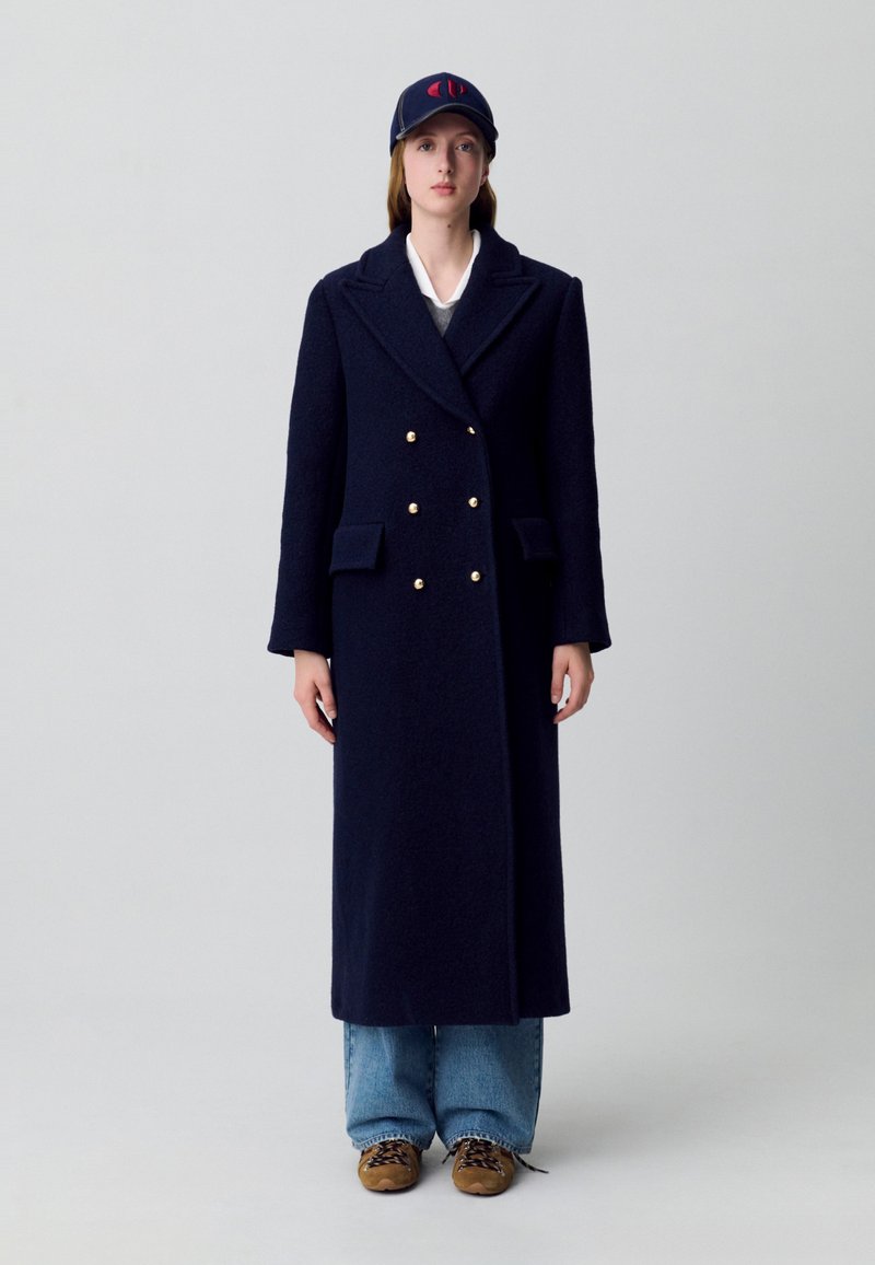 Cappotto lungo in lana blu navy con bottoni dorati doppi, colletto a notched, due tasche frontali, abbinato a un maglione grigio e jeans larghi.