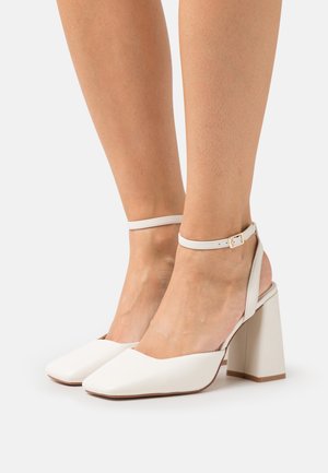 Chaussures à talons hauts blanches avec un bout carré, texture en cuir lisse, bride de cheville et talon bloc épais.