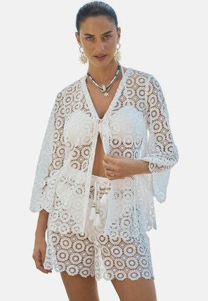 Femme portant un cardigan en dentelle crochet blanche et un short assorti, accessoirisée de boucles d'oreilles en forme de soleil et de colliers superposés.