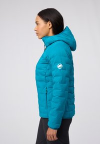 Turkos pufferjacka med dragkedja framtill, huva och sidofickor. Har en quiltad design och en rund logotyp på vänster ärm.