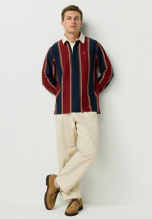 Camisa polo de manga larga con rayas rojas y azul marino, con cuello y un pequeño emblema. Combinada con pantalones beige y zapatos marrones.