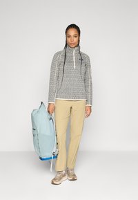 Pullover met een geometrisch zwart-wit patroon, beige broek, lichtblauwe rugzak en beige sneakers, staat tegen een effen achtergrond.