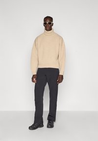 Beige gebreide coltrui, zwarte broek en zwarte sneakers. Model met lichtgekleurde zonnebril, staand tegen een effen achtergrond.
