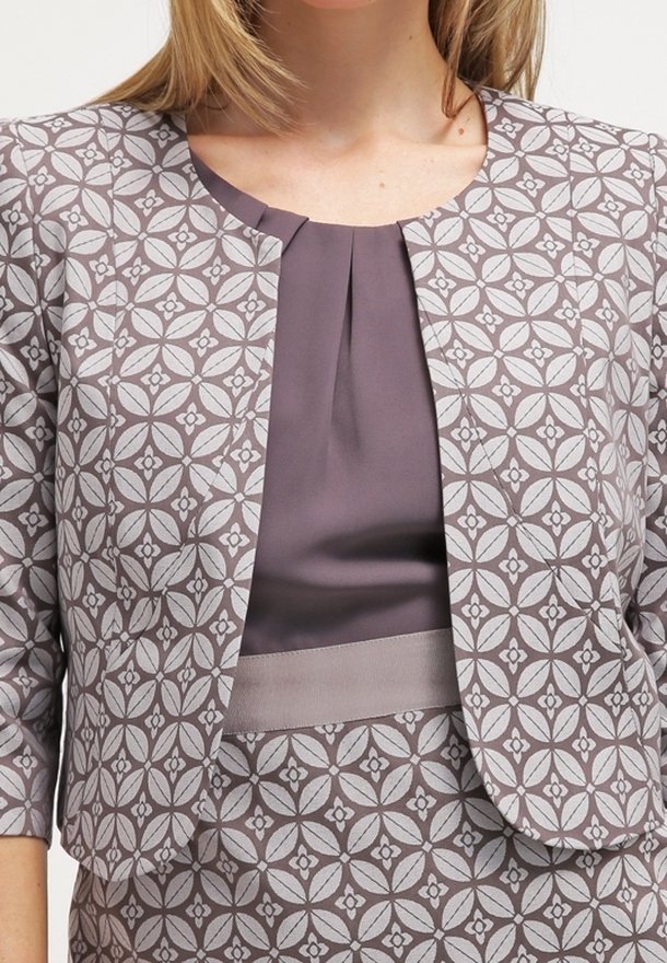 Femme portant une veste grise et blanche à motifs sur un chemisier plissé gris uni, assortie à une jupe à motifs similaires.