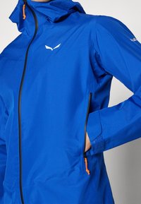 Blå vattentät jacka med huva, som har en frontdragkedja, orange drag och sidofickor. Slät textur med en logotyp på bröstet.