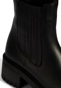 Zwarte leren enkelboot met verticale ribbels, stevige zool en een gladde textuur. Beschikt over een lage hak en zij-elastische panelen voor gemakkelijk aan- en uittrekken.
