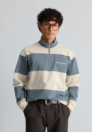 Junger Mann mit lockigem Haar und Brille trägt einen blau-beigen gestreiften Pullover mit Viertelreißverschluss und schwarze Hosen, die Hände in den Taschen.