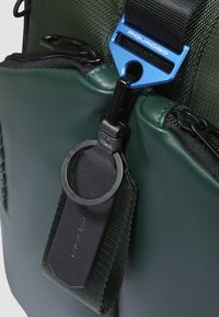 Borsa in pelle verde con dettagli in tessuto strutturato. Presenta una fibbia metallica blu e un attacco portachiavi nero per una maggiore funzionalità.