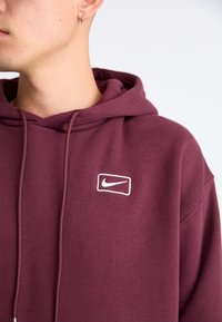 Maroon hoodie with a drawstring and a white embroidered Nike logo. Smooth fabric texture, relaxed fit, and a kangaroo pocket.