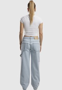 Kobieta z długim kucykiem stoi tyłem, ubrana w dopasowany biały crop top oraz luźne jasnoniebieskie dżinsy z tylnymi kieszeniami i beżową metką.