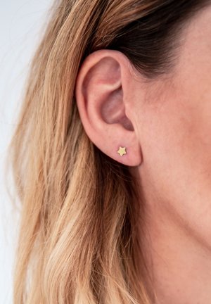 Goldfarbene Sternsteckerohrringe, die an einem Ohr getragen werden, mit einer glatten Oberfläche und minimalistischen Design in Form eines kleinen, fünfzackigen Sterns.