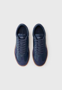 Marineblå joggesko laget av en kombinasjon av lær og semsket skinn. Rund tå, snøring og beige gummisåle. Lacoste-logo på tungen.