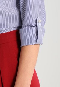 Blue and white striped shirt with rolled cuffs featuring a pearl button, paired with red high-waisted trousers, showing fabric texture.