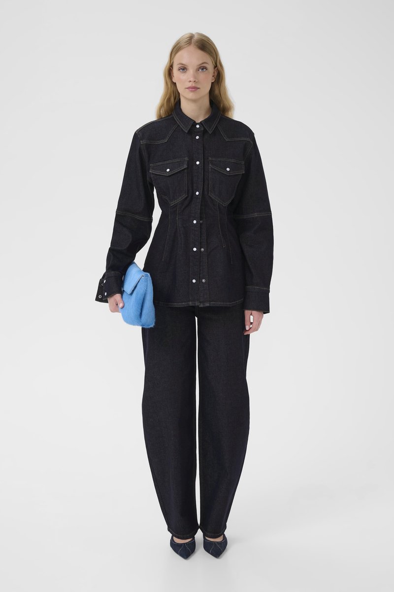 Dark denim shirt with two chest pockets, button closure, and contrasting stitching. Paired with wide-leg dark denim pants and blue clutch.