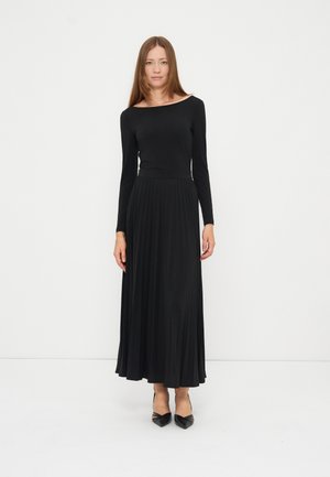 Femme aux longs cheveux raides portant une longue robe noire à manches longues et des talons noirs pointus, debout sur fond blanc.