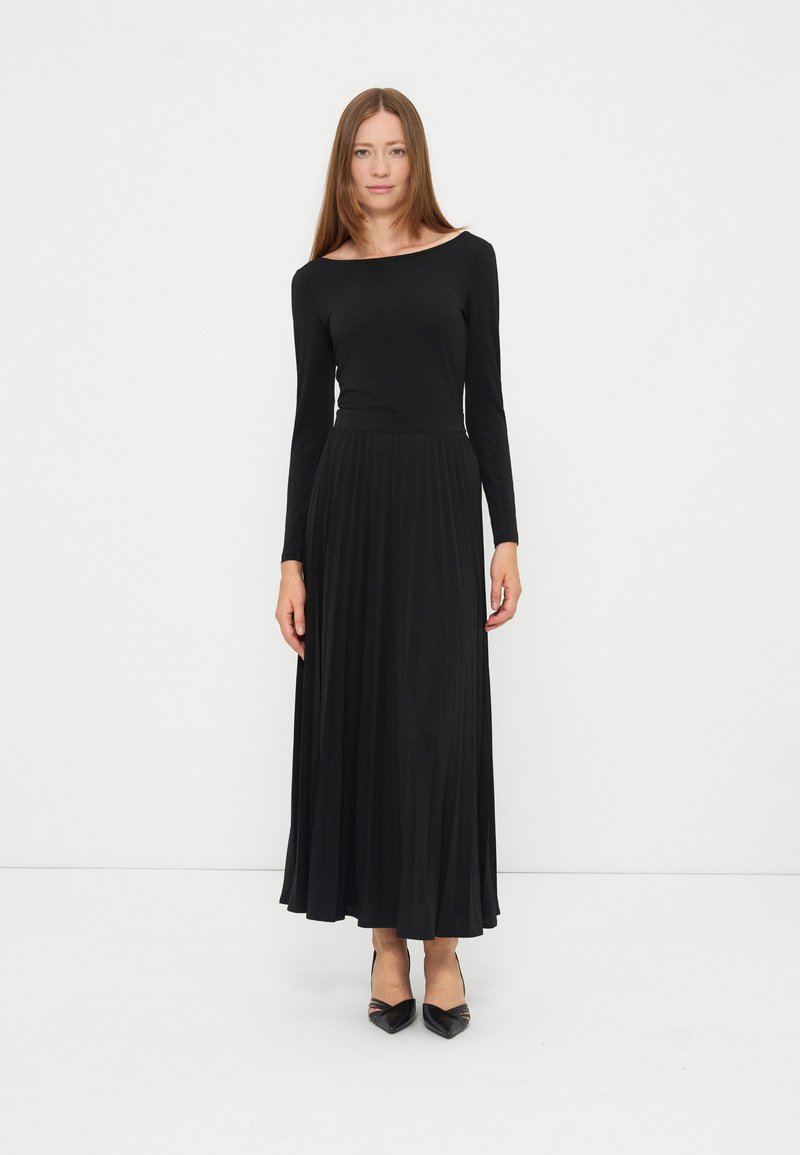 Femme aux longs cheveux raides portant une longue robe noire à manches longues et des talons noirs pointus, debout sur fond blanc.