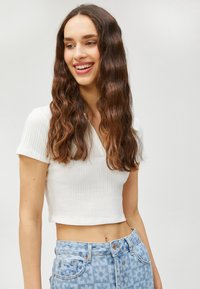 Weißes geripptes kurzes Oberteil mit kurzen Ärmeln, kombiniert mit hellblauen Jeans im Blumenmuster, die Taschen und gestickte Details aufweisen.