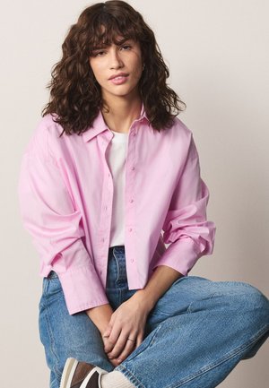 Femme aux cheveux bruns bouclés portant une chemise rose à boutons, un haut blanc, un jean bleu et des baskets marron, assise en tailleur sur un fond uni.
