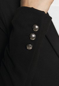 Černý strukturovaný textilní rukáv s třemi dekorativními stříbrnými knoflíky s jemnými vzory poblíž manžety.