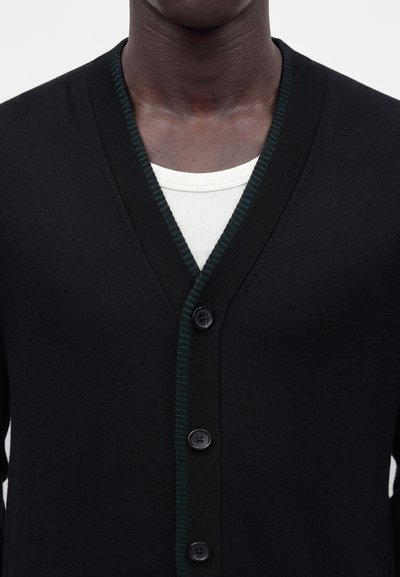 Homme portant un cardigan noir boutonné avec un bord vert foncé par-dessus une chemise blanche côtelée à col rond, présenté des épaules jusqu’au cou.