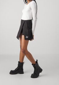 Jupe sportive noire avec fente sur le côté, associée à un haut blanc à manches longues. Bottines noires à lacets avec semelles épaisses. Logo Puma sur la jupe.