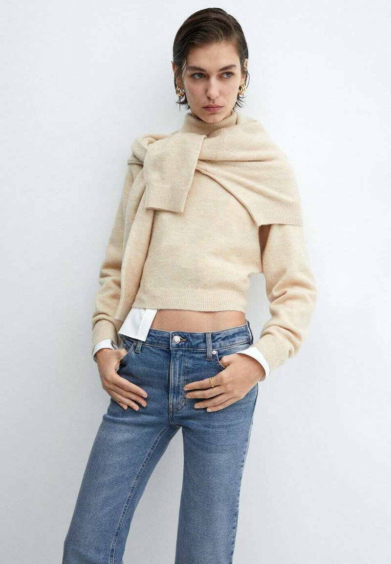 Beige cropped sweater met een wikkelstijl halslijn; gecombineerd met lichtblauwe high-waisted jeans. Soepele textuur, casual pasvorm en gelaagde look.