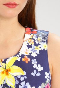 Blå blommig klänning med rundad hals, livfulla gula och orange blommor, strukturerad tyg och en figurnära silhuett.