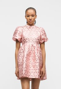 Γυναίκα που φοράει λαμπερό ροζ μίνι φόρεμα με φουσκωτά μανίκια και σχέδια καρδιών, στέκεται μπροστά σε απλό λευκό φόντο.