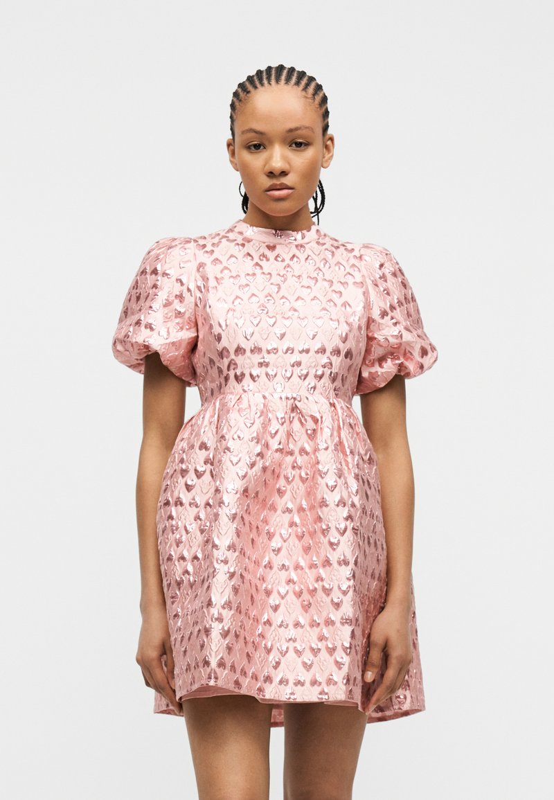 Γυναίκα που φοράει λαμπερό ροζ μίνι φόρεμα με φουσκωτά μανίκια και σχέδια καρδιών, στέκεται μπροστά σε απλό λευκό φόντο.