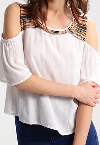 Top bianco con spalle scoperte, colletto decorato con perline colorate e texture, maniche corte e larghe, e una vestibilità rilassata. Tessuto morbido con una sensazione leggera.