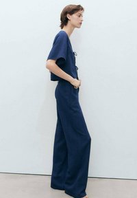 Temno modra skrajšana majica s kratkimi rokavi, z detajlom zavezovanja spredaj; v kombinaciji s širokimi hlačami iz teksturiranega materiala.