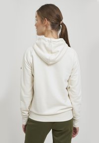 Hvit hettegenser med snorhette og ribbestrikkede mansjetter, laget av mykt stoff. Har en sydd logo-detall på venstre erm.