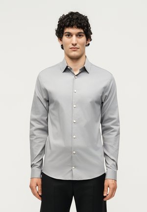 Jeune homme aux cheveux bouclés portant une chemise grise claire à manches longues boutonnée et un pantalon noir, debout devant un fond blanc uni.