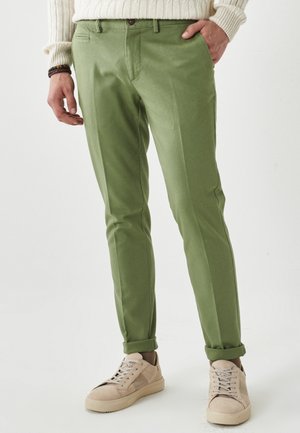 Mand iført grønne slim-fit bukser, beige sneakers og en cremefarvet kabelstrikket sweater, stående med den ene hånd i lommen.