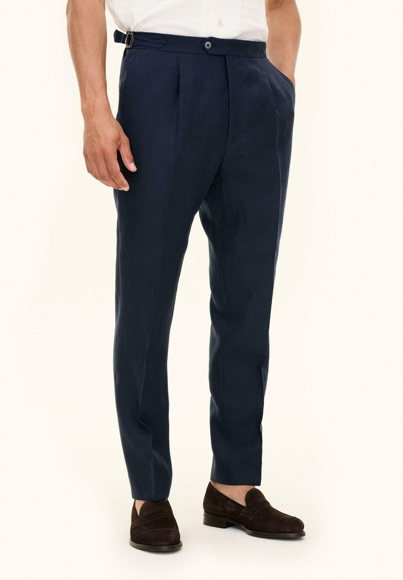 Mann trägt marineblaue, maßgeschneiderte Hose mit Seitenverstellern, weißes Hemd in die Hose gesteckt und dunkelbraune Wildleder-Loafer.