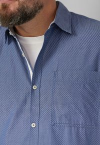 Camicia a pois blu realizzata in tessuto liscio. Presenta un colletto abbottonato, una tasca sul lato sinistro e dettagli in bottone bianco.