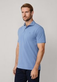 Gaiši zils polo krekls, kas izgatavots no mīksta auduma, ar norādītu apkakli, divām pogām un izšuvumu kreisajā krūtī. Īsas piedurknes.