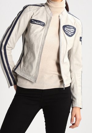 Person trägt eine hellfarbige Jacke mit Reißverschluss, dunklen Streifen an den Ärmeln und Aufnähern auf Brust und Arm, einen beigefarbenen gerippten Rollkragenpullover und schwarze Hose.