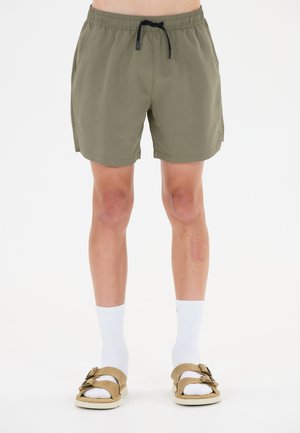 Person iført olivengrønne shorts, hvide crew-sokker og beige sandaler med dobbeltsnørre, vist fra taljen til fødderne mod hvid baggrund.