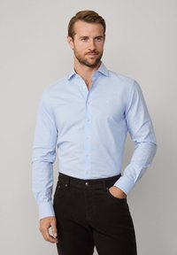 Camisa de botones de color azul claro, con mangas largas y collar, que presenta un pequeño logotipo en el pecho, combinada con pantalones oscuros.