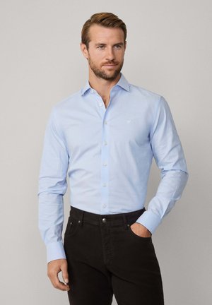 Chemise bleu clair à manches longues et col, avec un petit logo sur la poitrine, associée à un pantalon foncé.