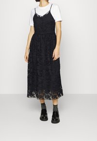 Robe midi en dentelle noire avec un motif floral et des bretelles spaghetti ajustables, superposée sur un t-shirt blanc, avec des chaussures noires.