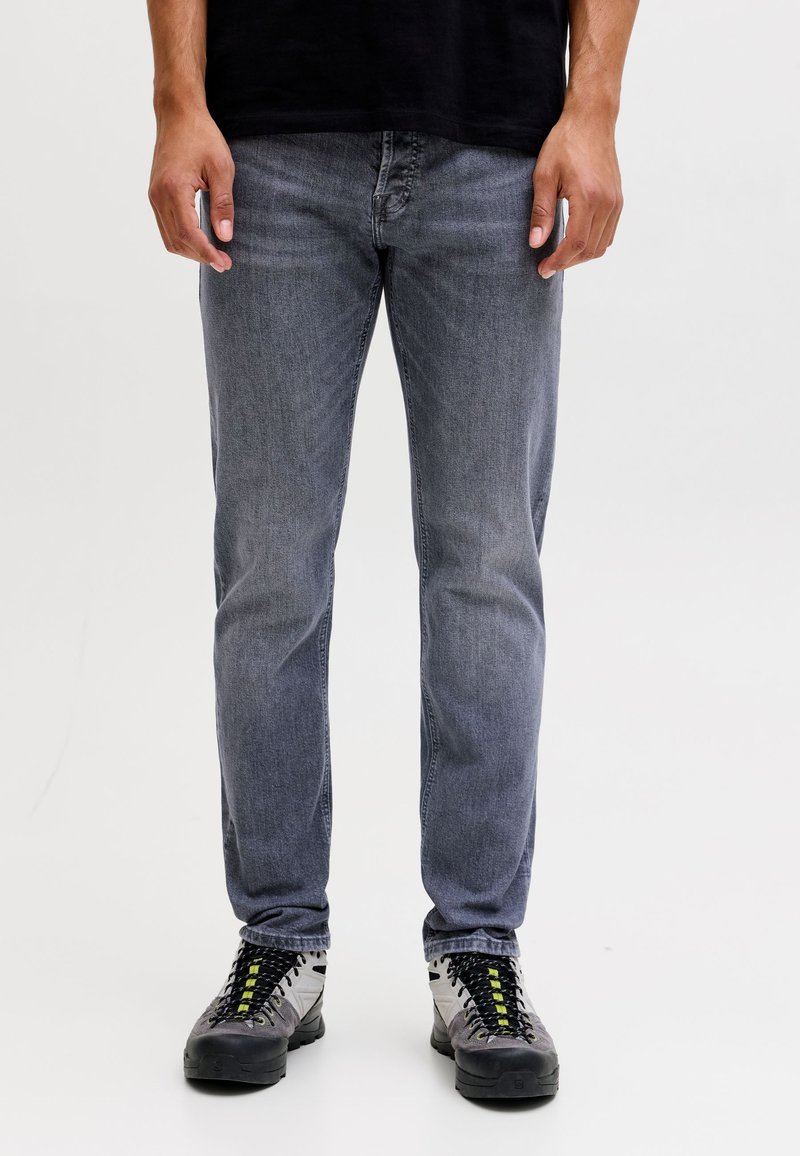 Mand stående i falmede grå jeans og hvide, grå og sorte vandresko med gule detaljer på en hvid baggrund.