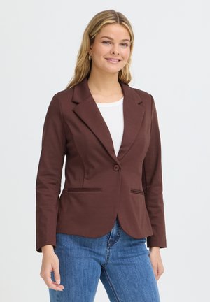 Jeune femme aux longs cheveux blonds, portant un blazer marron par-dessus un top blanc et un jean bleu, souriante sur un fond uni.