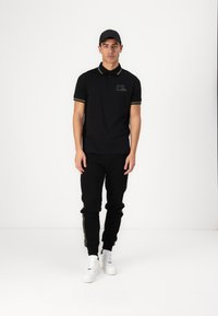 Schwarzes Polohemd mit gelben Akzenten, besticktem Logo, kombiniert mit schwarzen Jogginghosen mit gelben Seitenstreifen und weißen Sneakers.