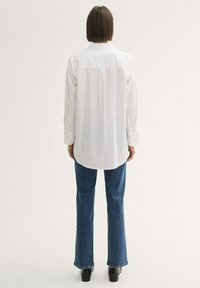 Weißes Langarmhemd mit geradem Schnitt, Rückenpasse und geknöpften Manschetten, kombiniert mit dunkelblauen Schlagjeans und schwarzen Ankle Boots.